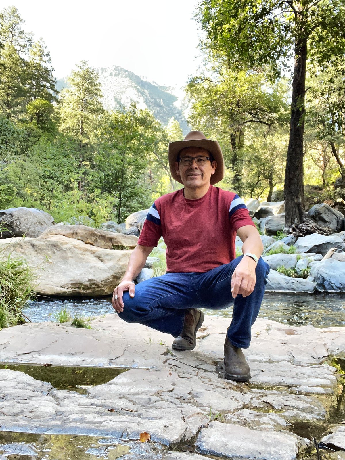 Tony Skrelunas in a brown felt hat, red shirt and jeans, crouching on flat sandstone ledges beside a shallow creek, with pools of water standing in the rock at his feet and pines and a forested ridge behind him.