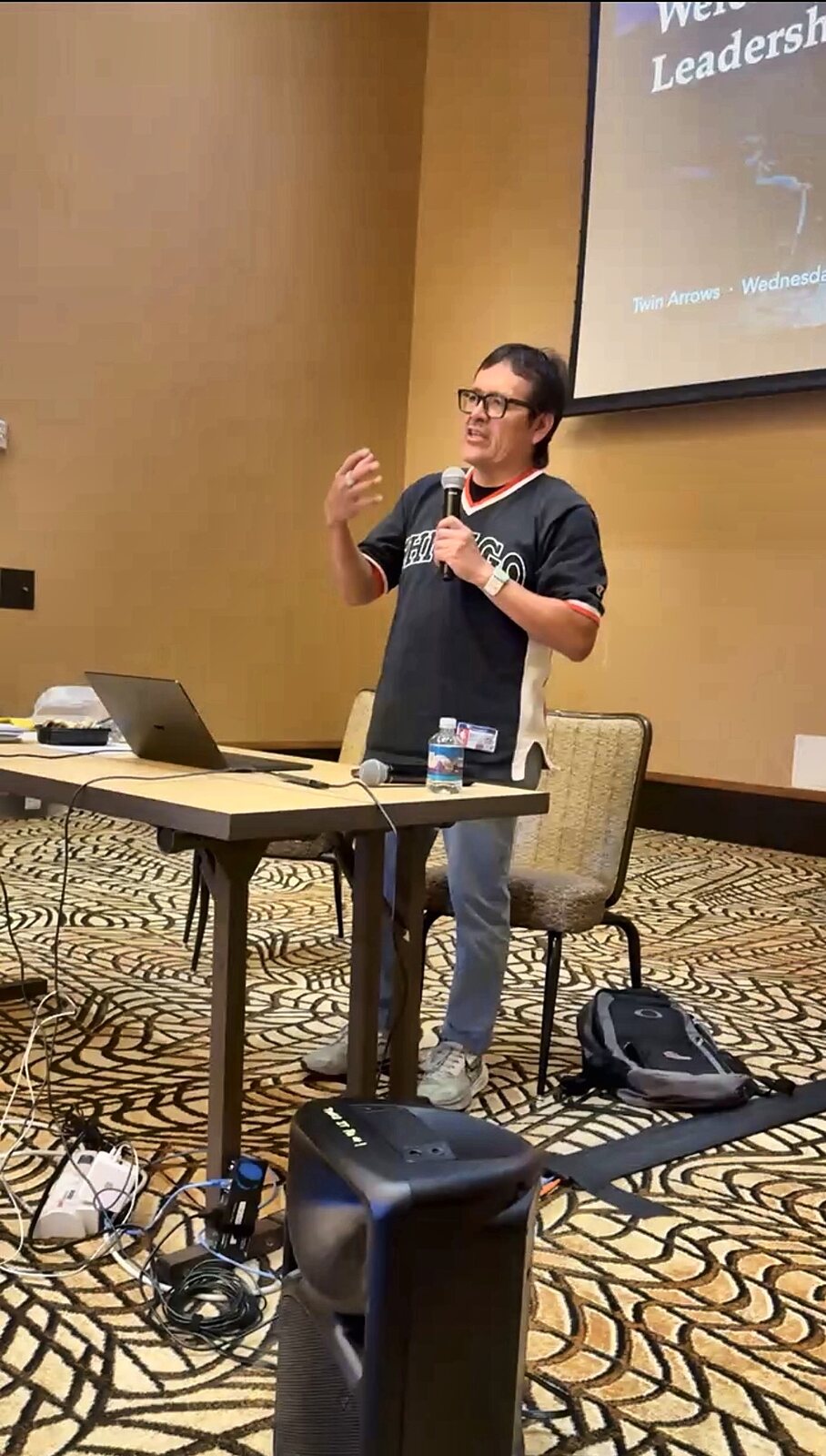 Tony Skrelūnas, microphone in hand, speaking at the front of the room at Twin Arrows, the welcome slide for the Hopi Leadership Session on the screen behind him