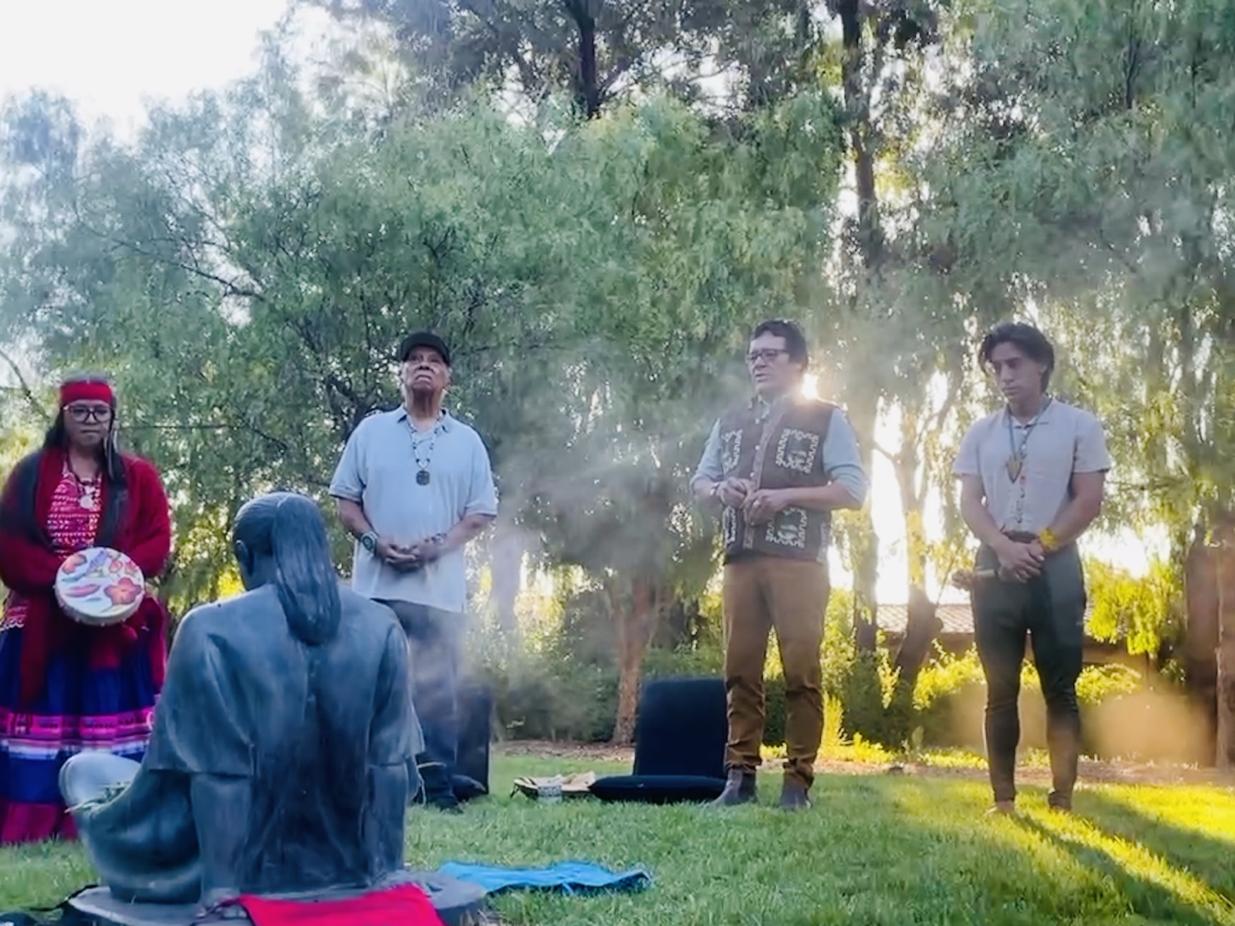 Tony Skrelūnas leading an outdoor ceremony circle with a hand drum and smudge